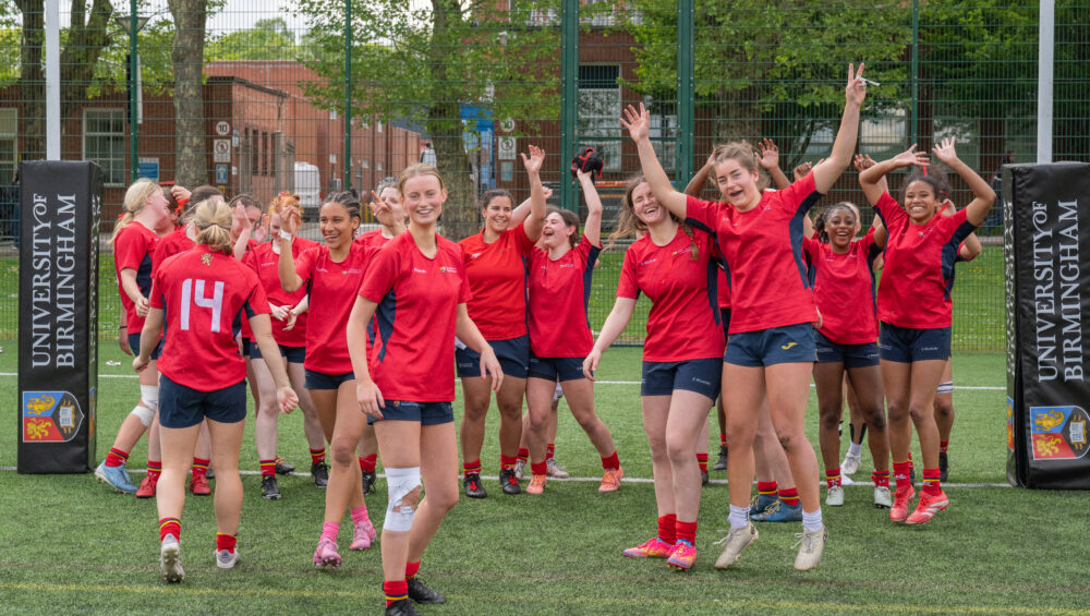 Womens Rugby team cheering after their win, standing in a large group, wearing their red University of Birmingham kit.