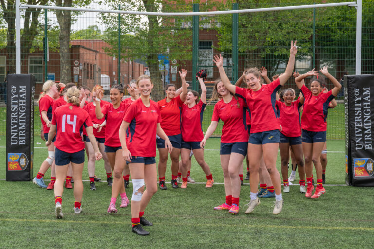 Womens Rugby team cheering after their win, standing in a large group, wearing their red University of Birmingham kit.