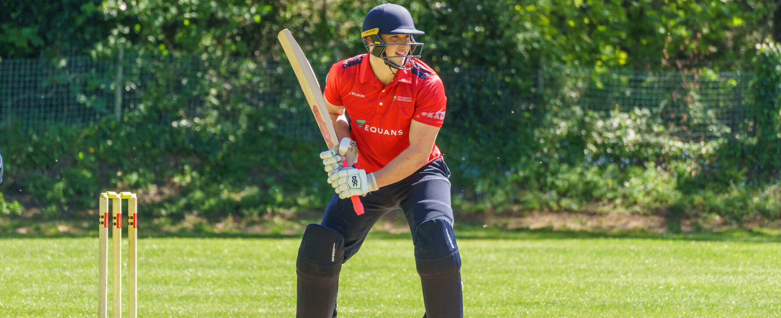 Male student holding cricket bat, wearing red University of Birmingham sport kit, along with cricket helmet.