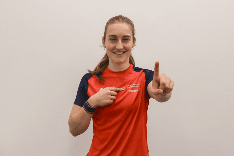 Evie Wood pointing at the UoB Sport & Fitness logo and putting one finger up.