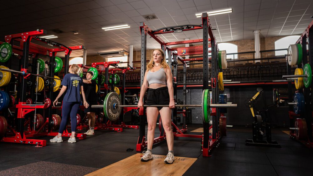Woman performing deadlifts in Tiverton gym with red squat racks with the university of birmingham logo behind her