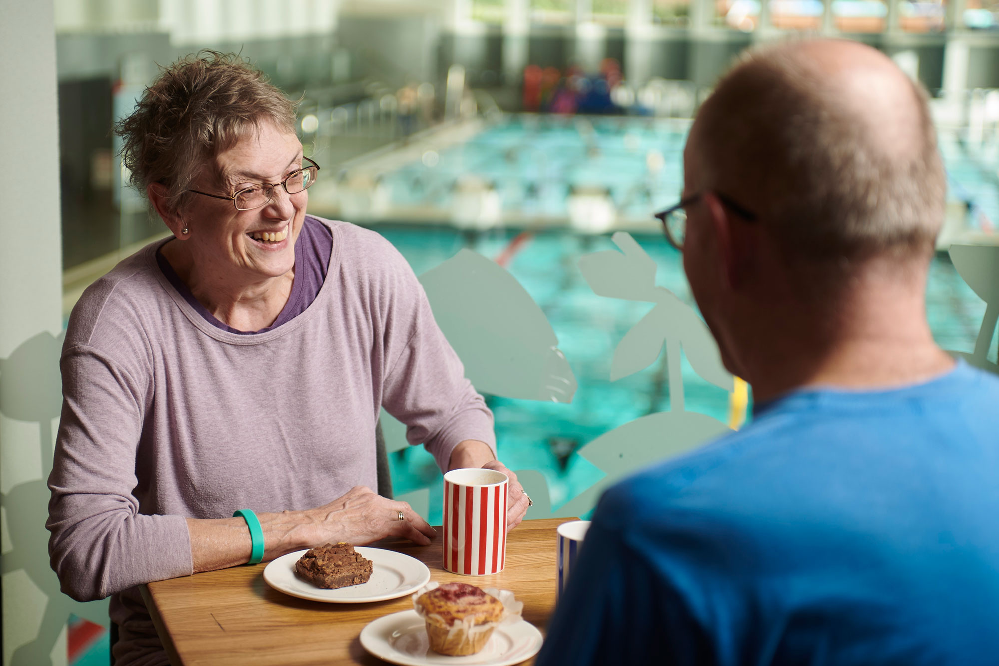 Two people sitting in Costa with a drink and a cake, there is a view of the pool behind them