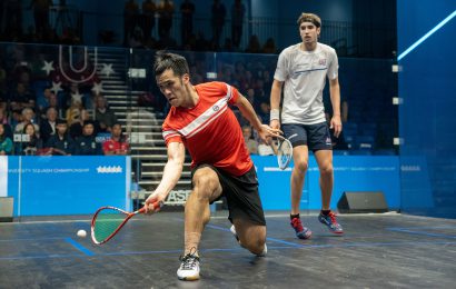 Two male squash players playing on an all glass showcourt in the Munrow Arena with a crowd behind them