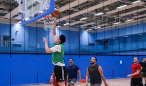 Basketball player wearing a green bib and glasses making a layup by jumping next to the hoop. 4 other players watch on from behind. Taken in munrow arena in sport and fitness gym, which has blue walls.
