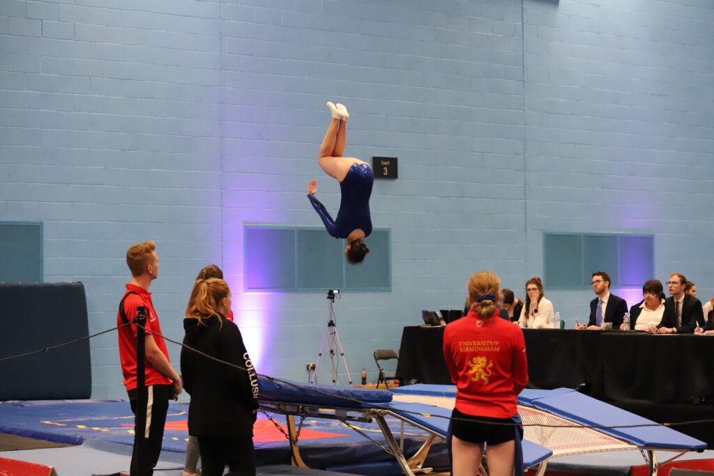 Female doing a flip mid-air after jumping on the trampoline, surrounded by coaches.