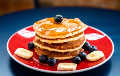 Stack of 4 pancakes drizzled with maple syrup, and topped with blueberries and bananas on a red plate.
