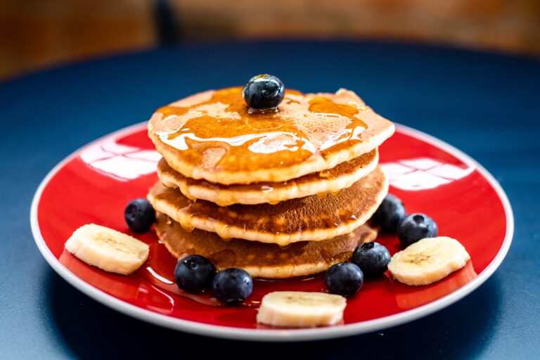 Stack of 4 pancakes drizzled with maple syrup, and topped with blueberries and bananas on a red plate.
