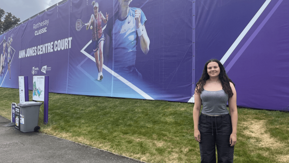 Georgia standing on the right hand side of large banner promoting Rothesay Classic Tennis event