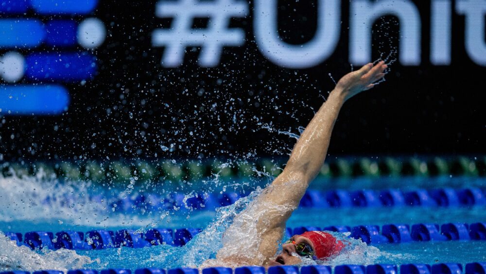 Scholar Ollie Morgan performing the backstroke, arm out of the water wearing a red hat.