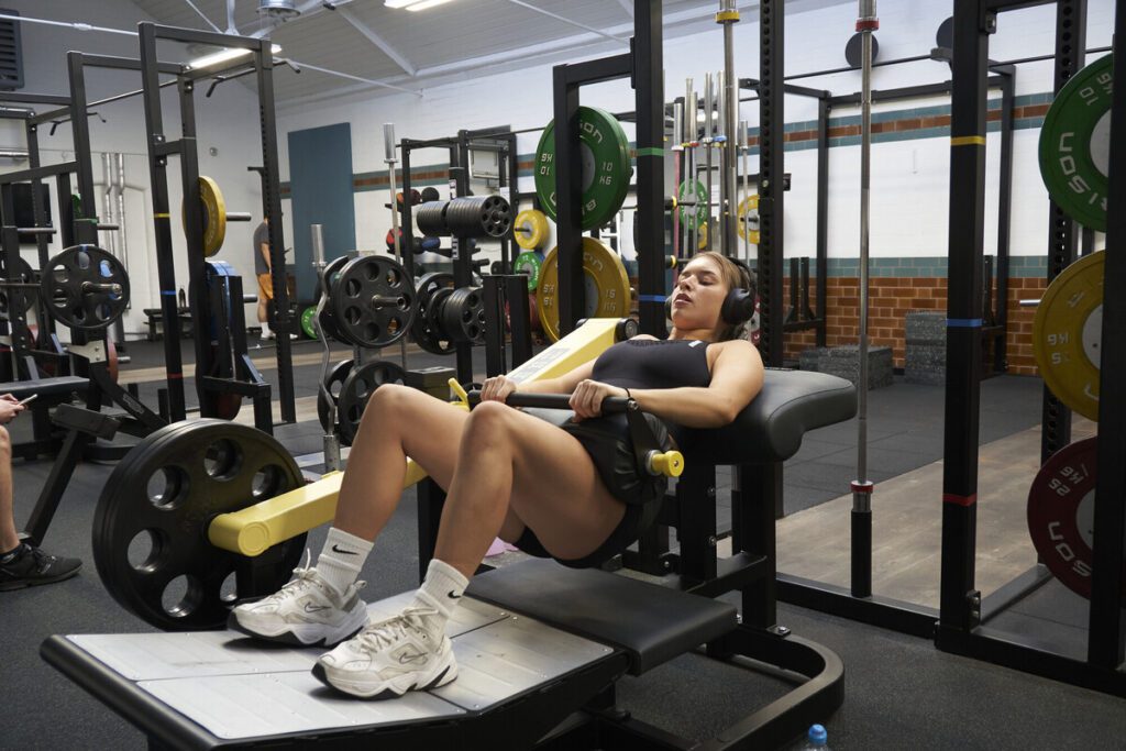 A member of Tiverton Gym, lying on and working out using the hip thrust machine. Squat racks are positioned behind her.