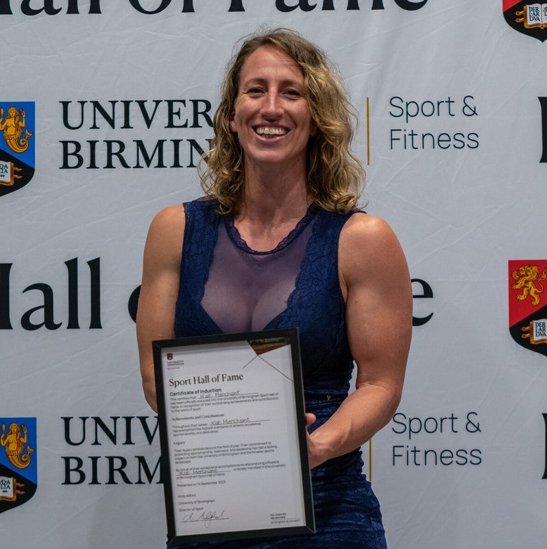 Kat Merchant with her Hall of Fame Award, standing in front of the Sport of Fitness/Hall of Fame sign.