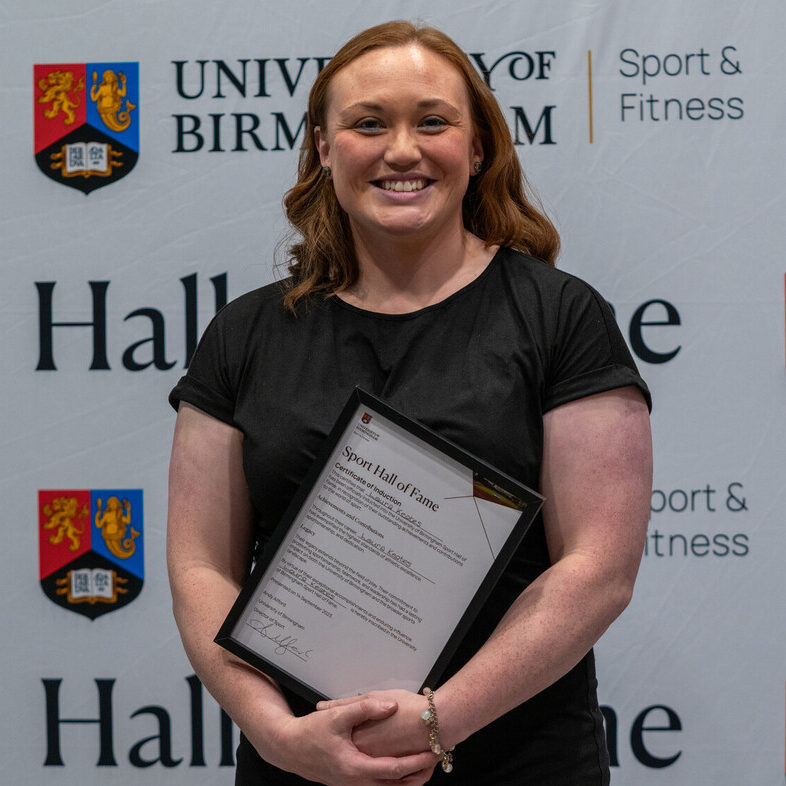 Laura Keates holding her Sport Hall of Fame award in front of the University of Birmingham Sport & Fitness backdrop.