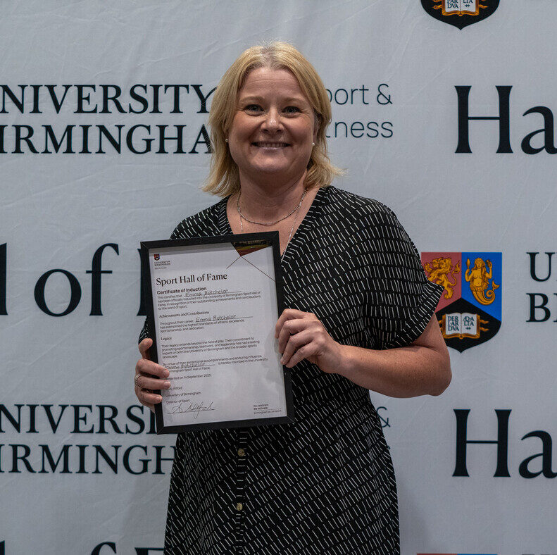 Emma Batchelor holding her Sport Hall of Fame award in front of the University of Birmingham Sport & Fitness backdrop.