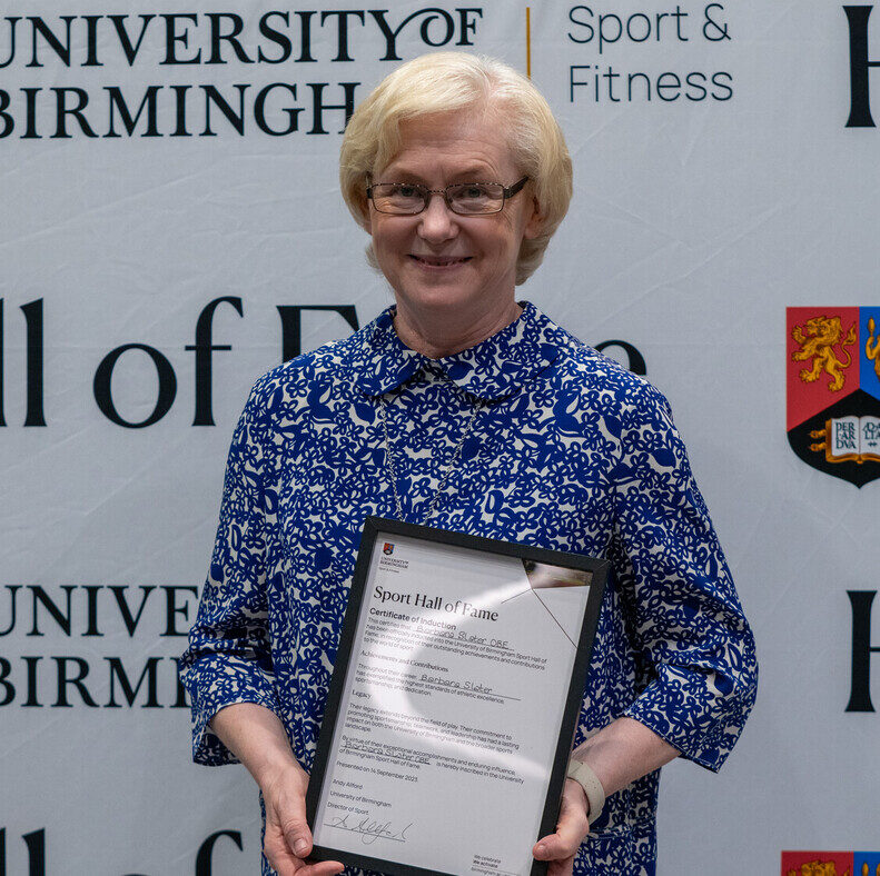 Barbara Slater holding her Sport Hall of Fame award in front of the University of Birmingham Sport & Fitness backdrop.