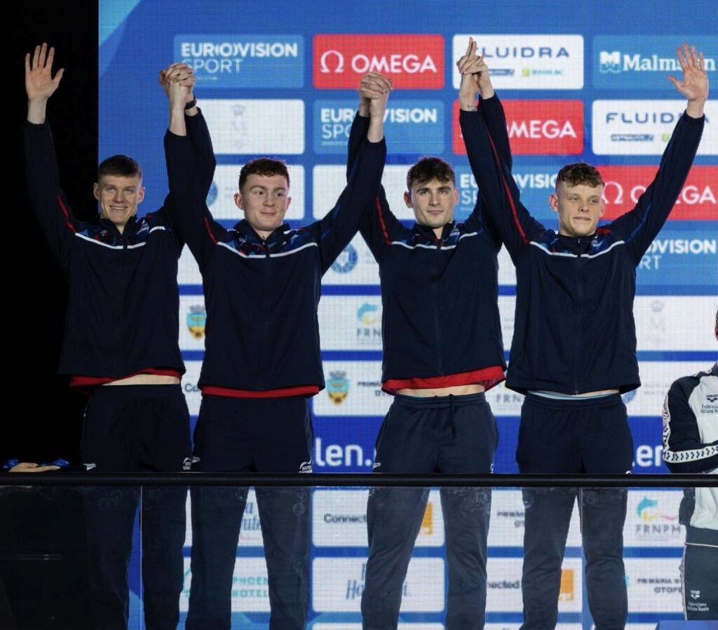 Four swimming athletes celebrating on a podium, holding hands in the air.