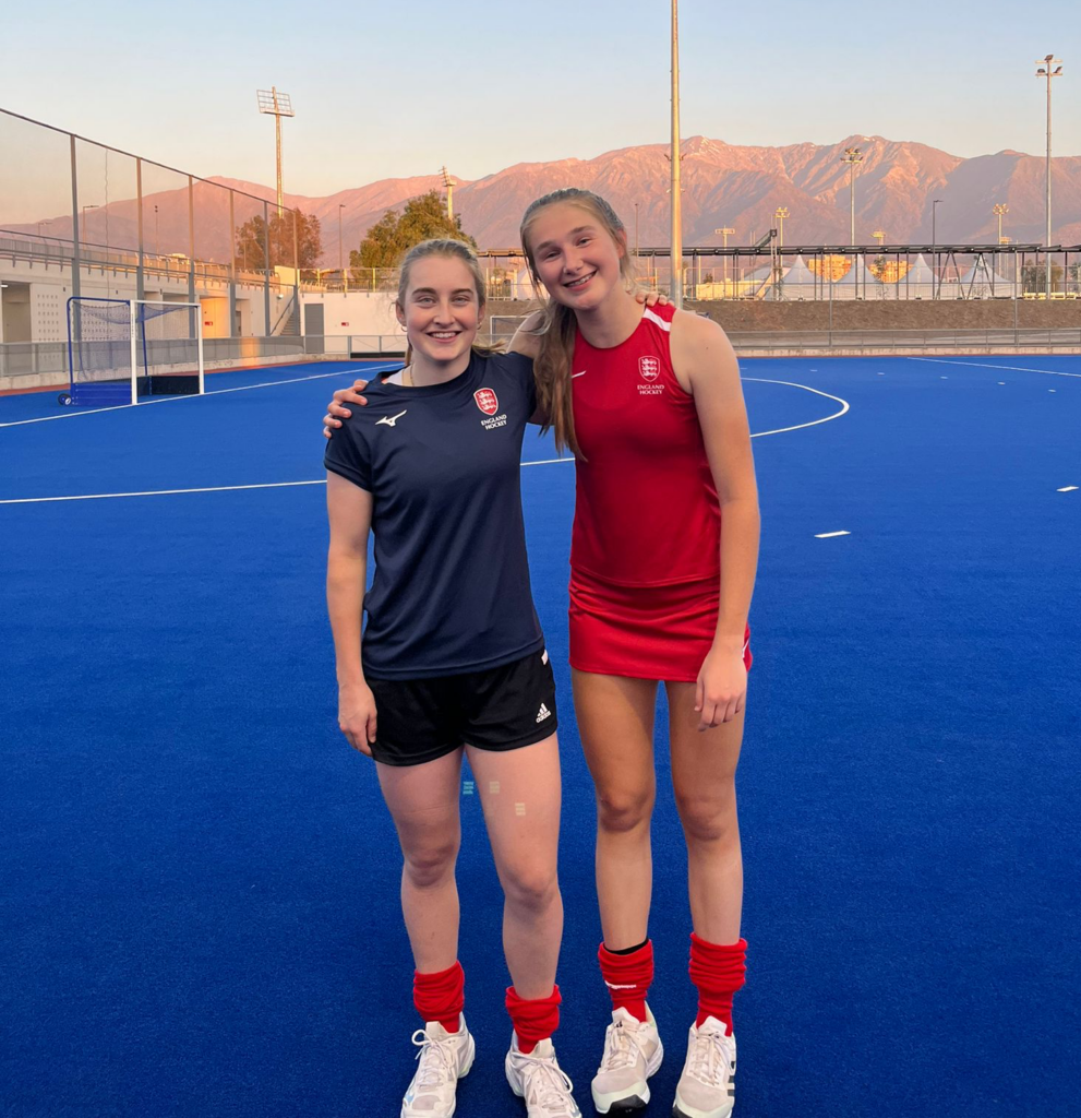 Evie Wood and Alice Atkinson stood on the hockey pitch hugging eachother and smiling.
