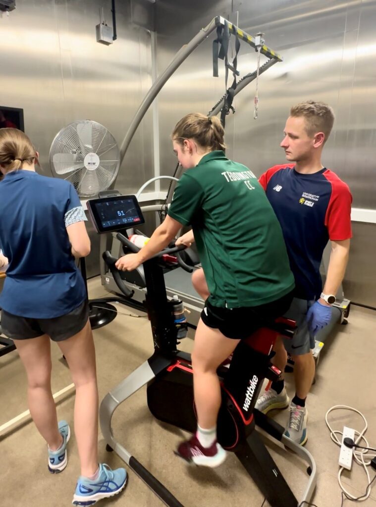 Evie Wood workin in the heat chamber, on a bike, with a member of the performance team supporting her.