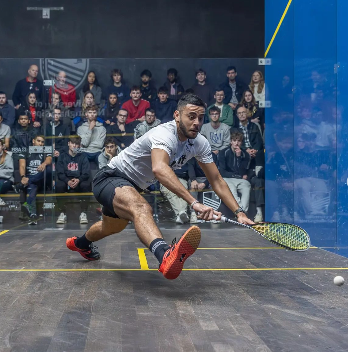 A squash player in a glass-back squash court bending to reach for the squash with the racquet.
