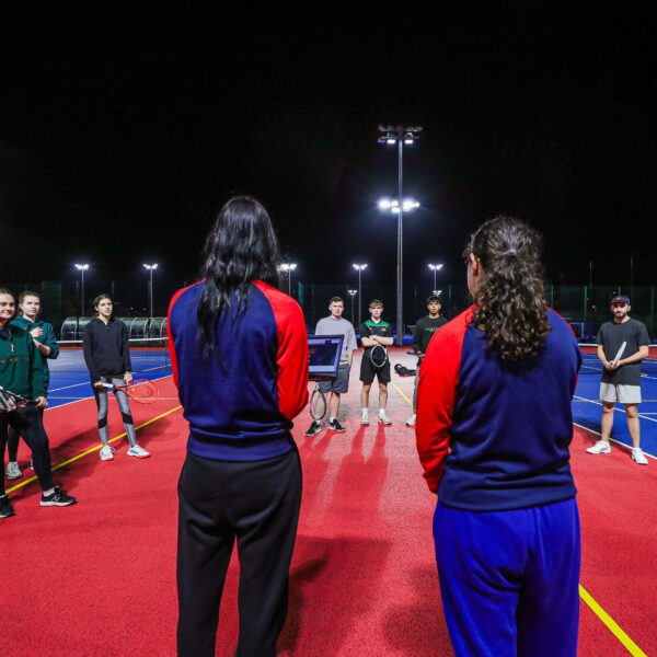 Two team leaders, dressed in uniform, holding a laptop and talking to a group of people on the tennis courts.