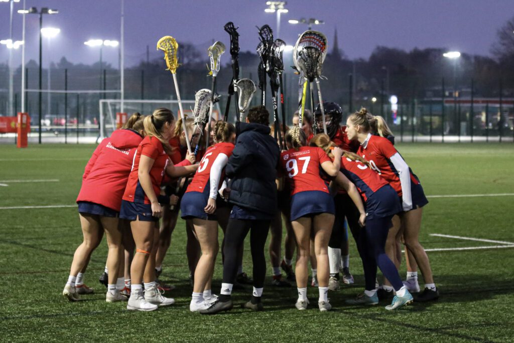 Group of lacrosse players in a huddle with lacrosse sticks up in the air.