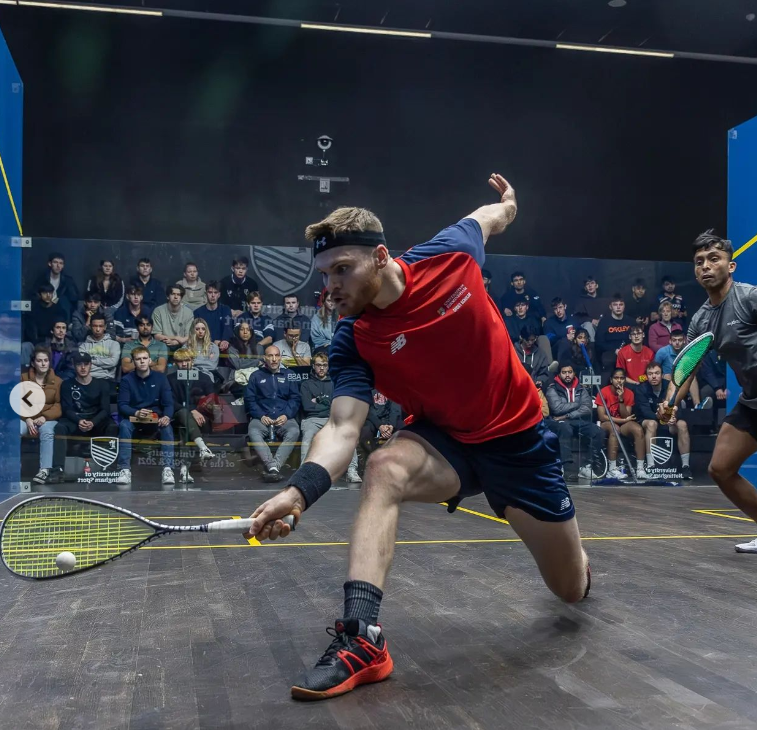 Will Salter in a glass-back squash court bending to reach for the squash with the racquet.