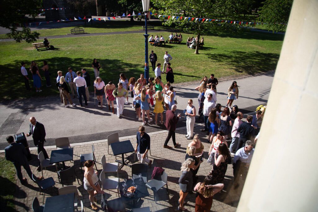 A group of students waiting outside in he sun on a grass area.