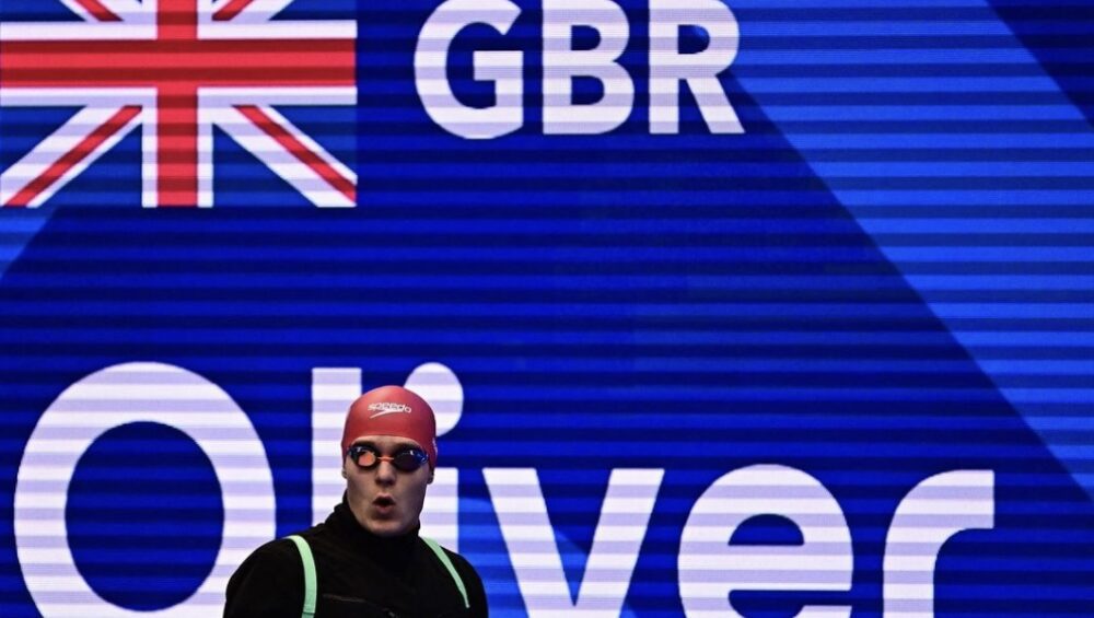 Oliver Morgan standing in front of a Great Britain sign wearing a swim cap and goggles.
