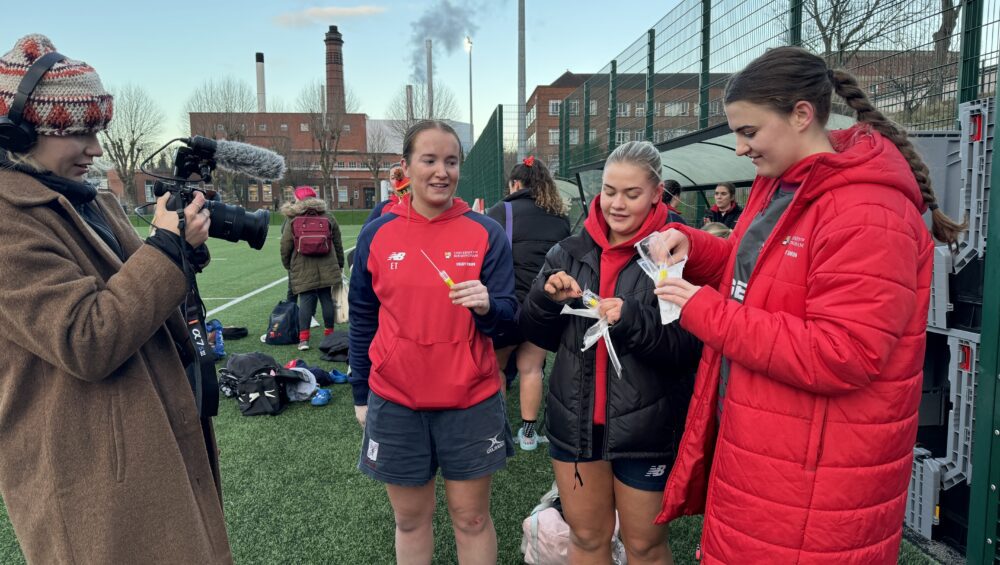 Three women from Rugby team taking diagnostic swab out of packaging, with BBC videographer filming.