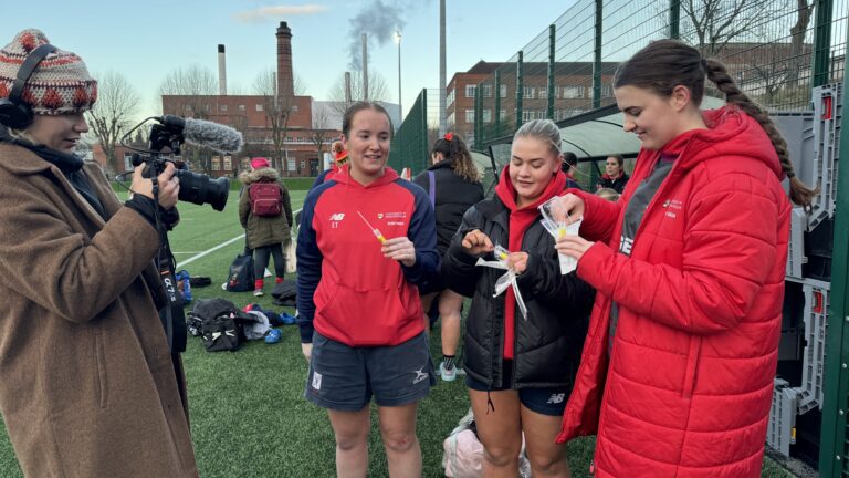 Three women from Rugby team taking diagnostic swab out of packaging, with BBC videographer filming
