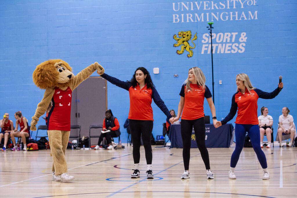 Three members of the netball club holding hands and preparing to Mexican wave with Rory the Lion.