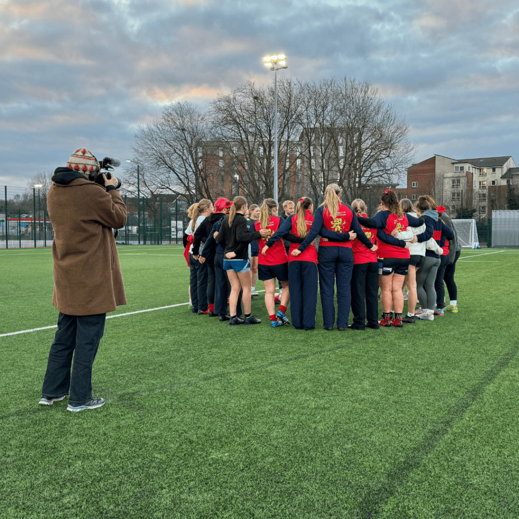 Women's Rugby team huddled on the pitch with camera.