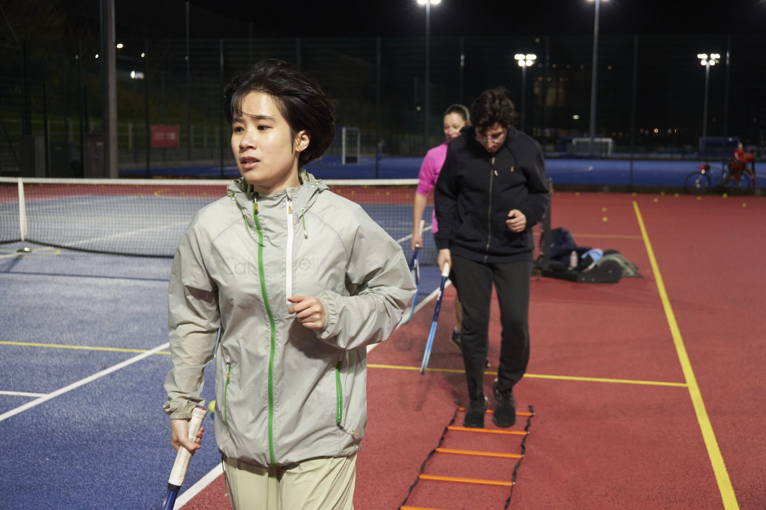 Two people running on the tennis courts, through a ladder on the floor holding tennis rackets in their hands.