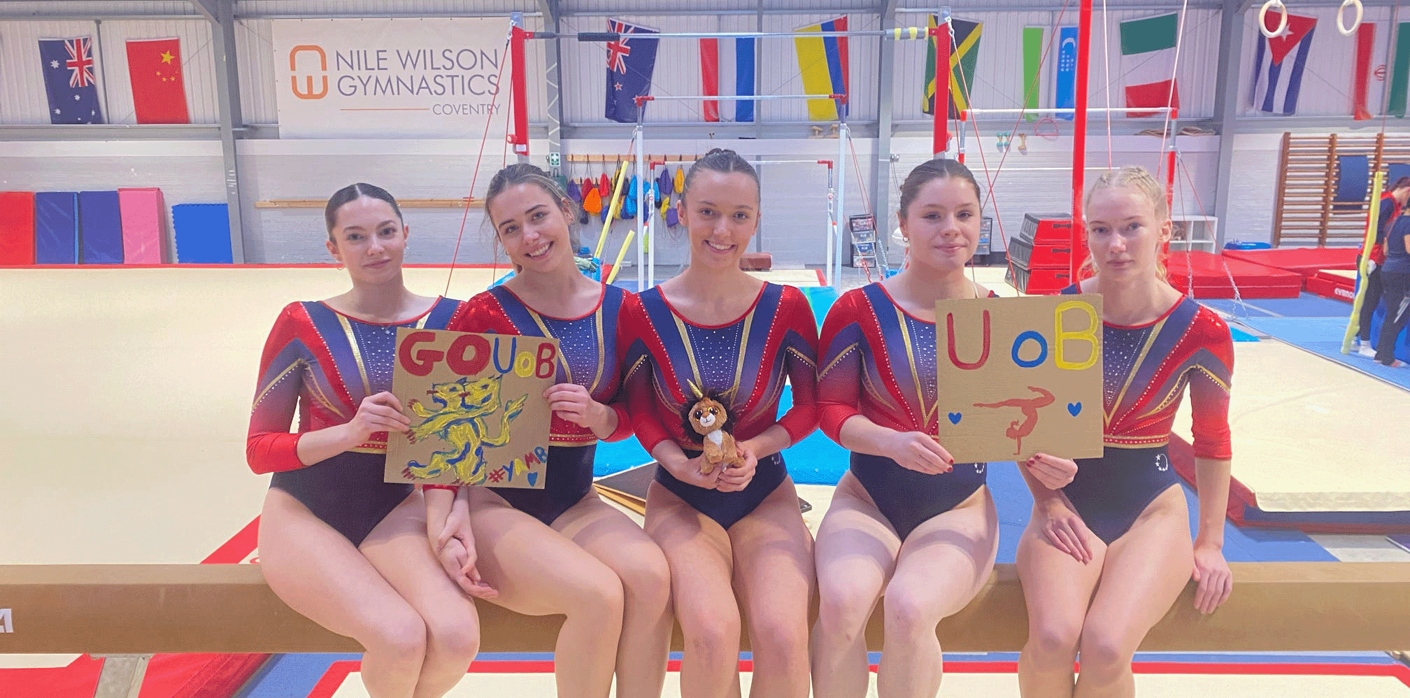 Gymnastics group sat together on a beam holding signs and smiling