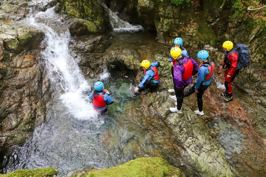 Students taking part in gorge walking, with one member of the group within the water and others on top of the rocks.