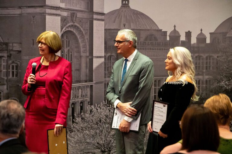 Three individuals standing after collecting their awards, listening to the speaker - holding a microphone and their certificates.