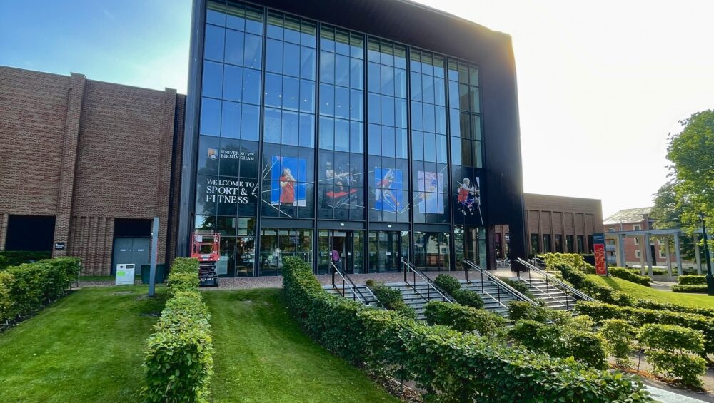 Exterior of University of Birmingham Sport & Fitness in Edgbaston. The sun shines over the building and decorative hedges at the entrance to the building.