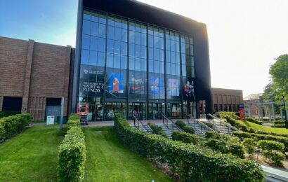 Exterior of University of Birmingham Sport & Fitness in Edgbaston. The sun shines over the building and decorative hedges at the entrance to the building.
