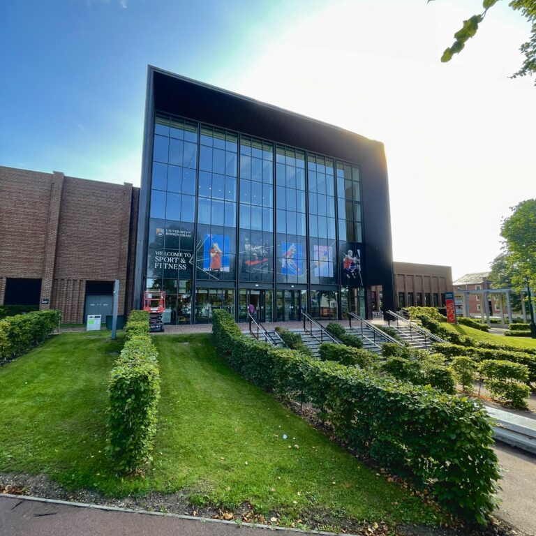 Exterior of University of Birmingham Sport & Fitness in Edgbaston. The sun shines over the building and decorative hedges at the entrance to the building.